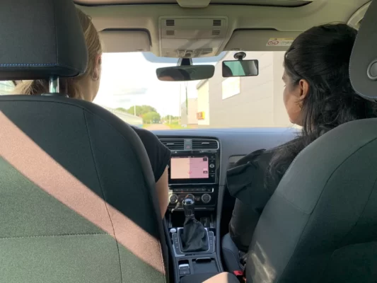 Foto vanuit de binnenkant van een lesauto vanuit de achterbank waarbij een dame bezig is met een rijles met daarnaast de rijinstructeur.